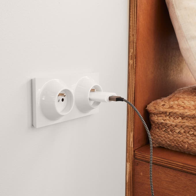 Photo Leroy Merlin d'une prise électrique blanche sur un mur blanc avec un câble branché. 
