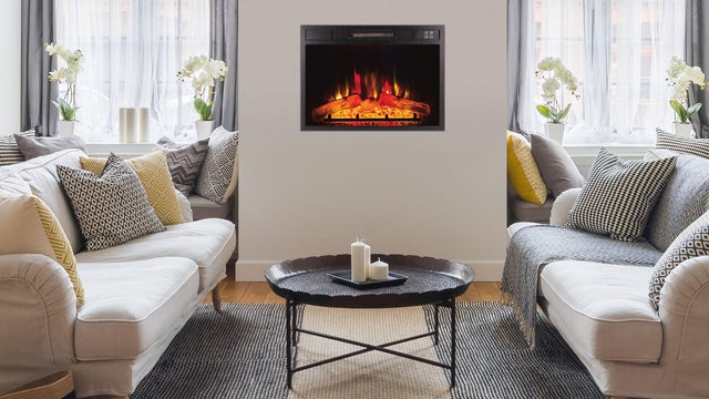 Sala de estar aquecida com salamandra elétrica na parede creme. Sala com dois sofás cremes e almofadas com padrão amarelo e preto. Mesa de centro preta. Cortinados cinzentos e brancos, flores. 