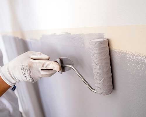 pessoa a utilizar um rolo de tinta com tinta cinzenta para pintar um rodapé na parede e a utilizar luvas de proteção.