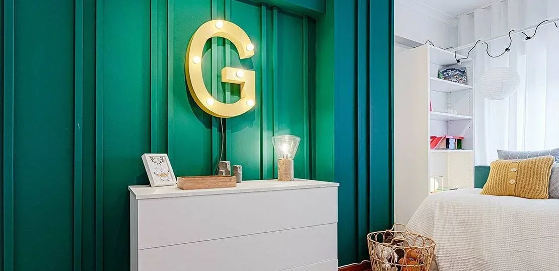 Quarto de criança com uma parede pintada a verde escuro com ripas de madeira coladas e um G luminoso 
