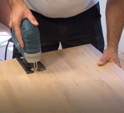 Duas mãos a cortar um tampo de madeira de cozinha com uma serra tico-tico