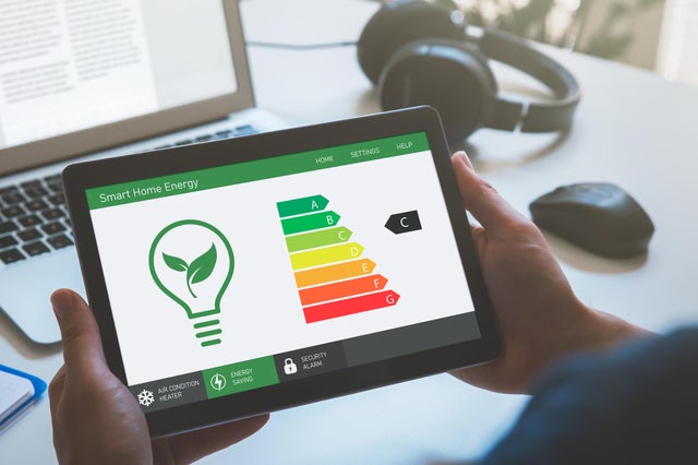 Mężczyzna trzyma w dłoniach tablet, na którym wyświetla się grafika z żarówką i klasami energetycznymi, w tle laptop i sluchawki