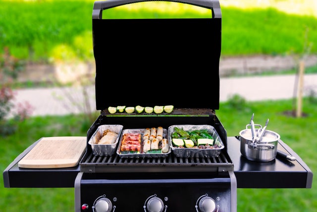 Otwarty grill gazowy z tackami pełnymi warzyw i mięsa.