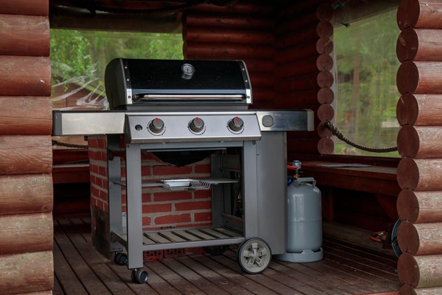 Grill gazowy na kółkach w drewnianej altanie z butlą gazową obok.