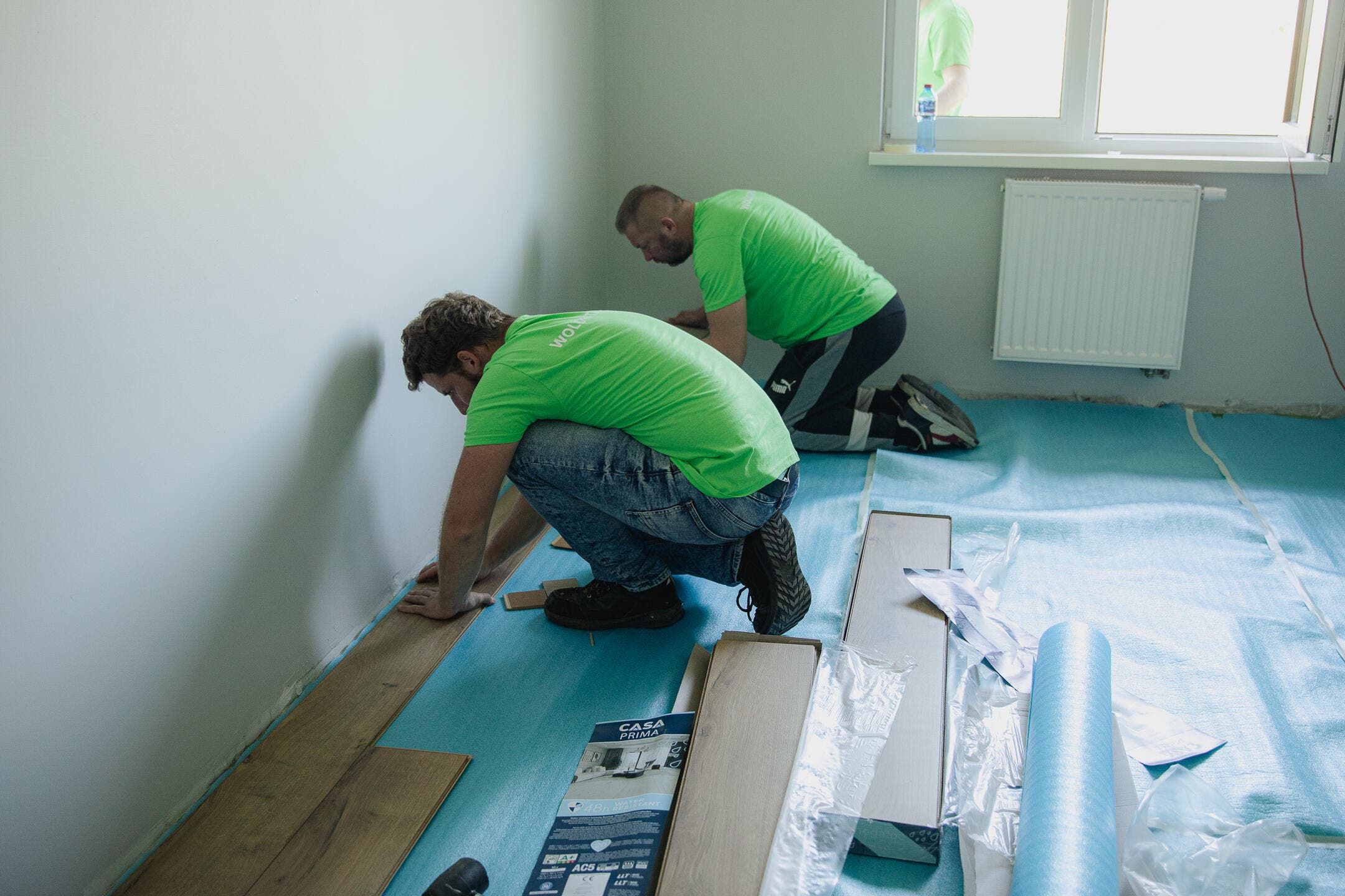 zdjęcie dwóch wolontariuszy układających piankę pod panele w remontowanym pokoju ośrodka