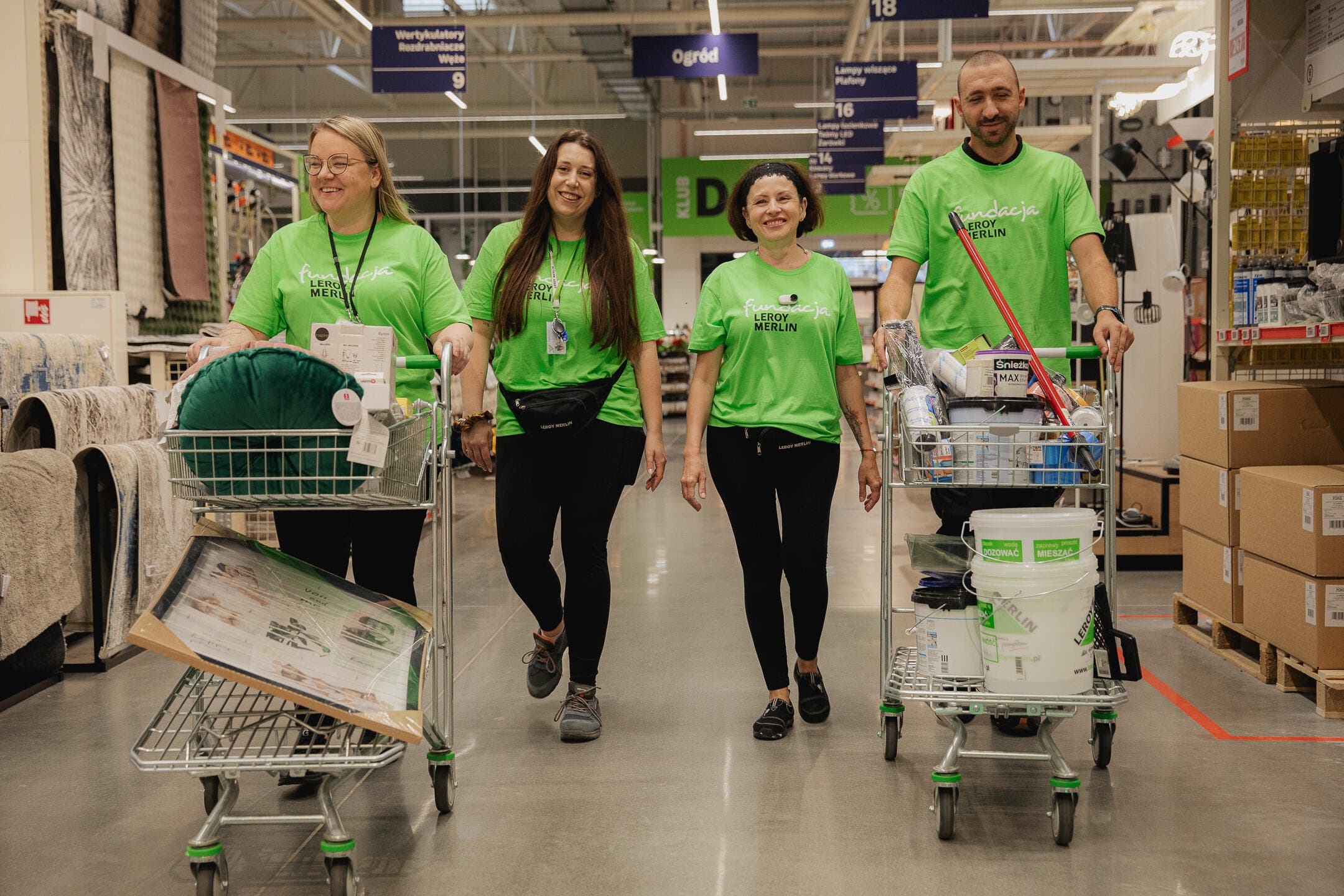 Uśmiechnięci Wolontariusze w robiący zakupy sklepie, przed sobą mają wózki sklepowe wypełnione artykułami potrzebnymi do remontu