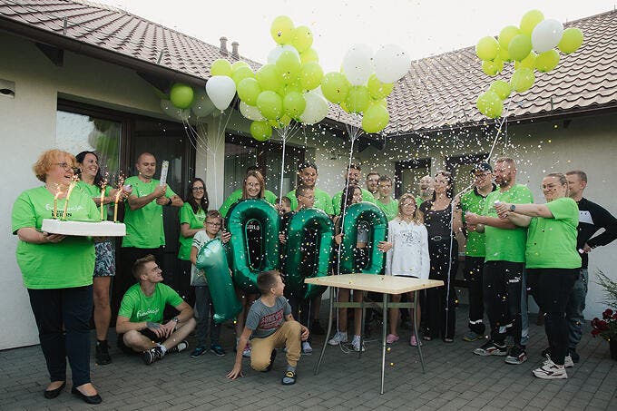 zdjęcie wolontariuszy i dzieci przed budynkiem ośrodka. Dzieci trzymają po kolei balony tworzące liczbę tysiąc. Jedna z osób wystrzeliła z racy konfetti.