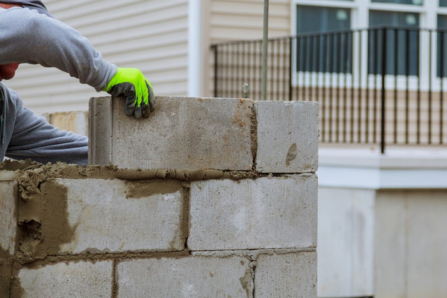 Układanie bloczków betonowych fundamentowych na zaprawie przez pracownika w rękawicach ochronnych.