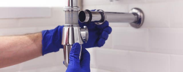 Montaż chromowanego syfonu umywalkowego przez hydraulika w rękawiczkach podczas remontu łazienki.