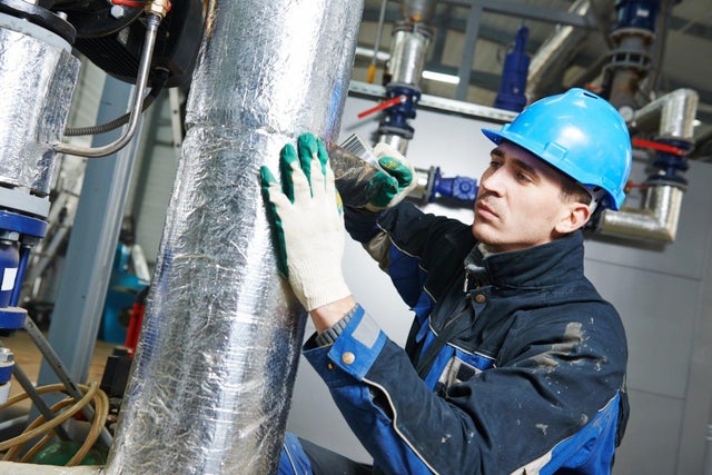 Pracownik techniczny montujący izolację na rurach instalacyjnych w pomieszczeniu przemysłowym.