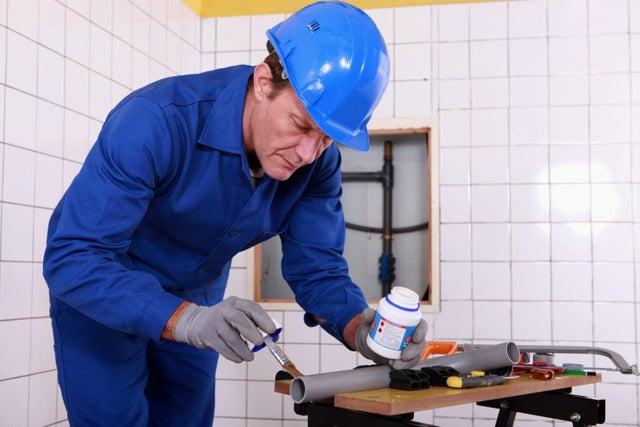 Hydraulik w niebieskim kombinezonie i kasku ochronnym nakładający pędzlem klej na szarą rurę PVC podczas montażu instalacji kanalizacyjnej w łazience.