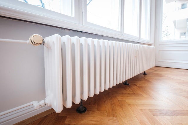 Biały żeliwny grzejnik członowy z głowicą termostatyczną zamontowany pod oknem na parkiecie.