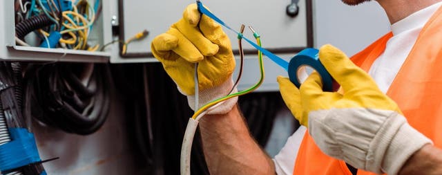 Elektryk w żółtych rękawicach roboczych owija niebieską taśmą izolacyjną przewód elektryczny przy skrzynce.