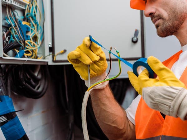 Elektryk w żółtych rękawicach roboczych owija niebieską taśmą izolacyjną przewód elektryczny przy skrzynce.