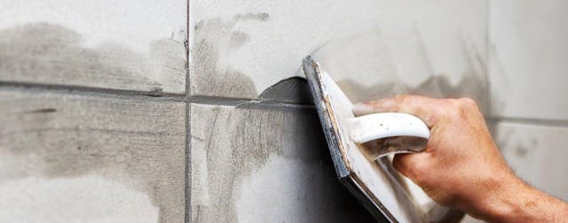 Osoba wcierająca szarą fugę między płytki ceramiczne na ścianie przy użyciu gumowej pacy podczas prac wykończeniowych w łazience.
