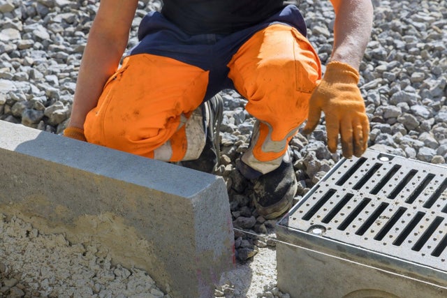 Pracownik budowlany w pomarańczowych spodniach i rękawicach montujący metalową kratę odwodnienia liniowego przy betonowym krawężniku na placu budowy.