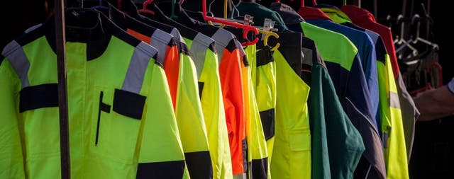 Kurtki i kamizelki ostrzegawcze z odblaskowymi pasami w kolorach żółtym i pomarańczowym wiszące na wieszaku w sklepie z odzieżą roboczą.