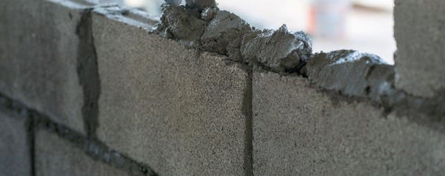 Murarz w rękawiczkach nakładający zaprawę murarską kielnią na bloczki betonowe podczas budowy ściany.
