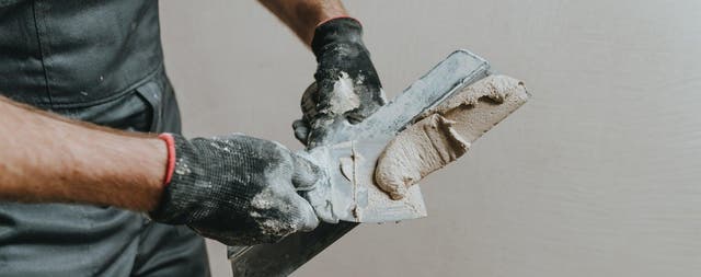 Fachowiec w czarnych rękawicach roboczych przekłada szpachlą gipsową gładź na szerszą pacę metalową.