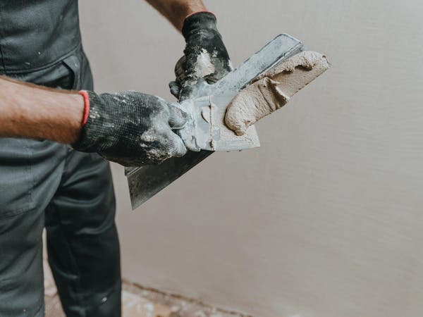 Fachowiec w czarnych rękawicach roboczych przekłada szpachlą gipsową gładź na szerszą pacę metalową.
