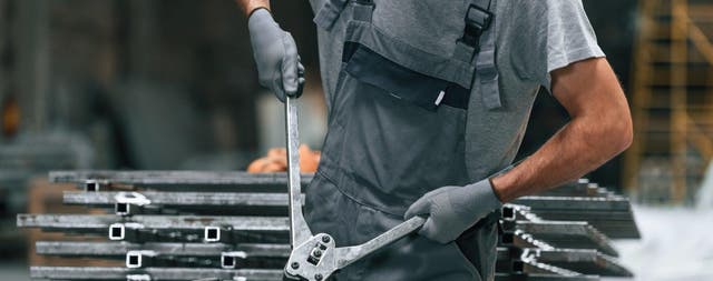 Pracownik w szarym ogrodniczkowym stroju przecina metalowe profile ręcznymi nożycami w warsztacie.