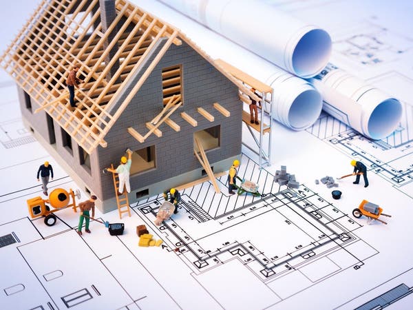 Miniaturowy model domu w trakcie budowy z figurkami robotników i rozwiniętymi planami architektonicznymi na białym tle.