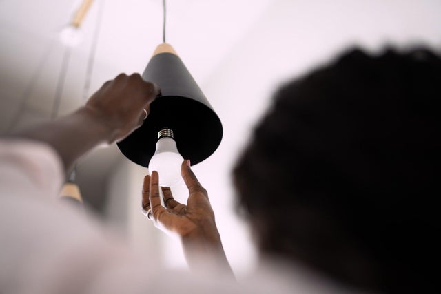Dłonie kobiety wkręcają energooszczędną żarówkę LED do czarnej lampy wiszącej pod sufitem.