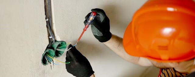 Elektryk w pomarańczowym kasku sprawdza przewody elektryczne w ścianie, używając czerwonego próbnika napięcia.