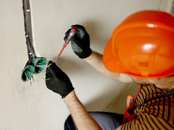 Elektryk w pomarańczowym kasku sprawdza przewody elektryczne w ścianie, używając czerwonego próbnika napięcia.