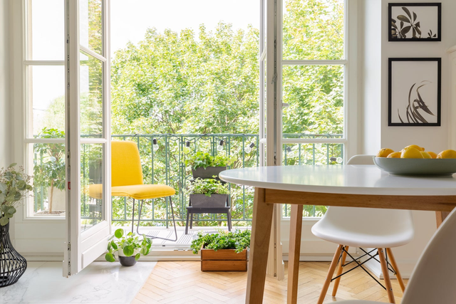 Otwarte białe drzwi balkonowe w salonie z jadalnią prowadzące na zielony balkon z roślinami.