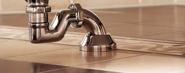 Chromowany syfon umywalkowy zamontowany pod białą umywalką na beżowych płytkach ceramicznych w łazience.