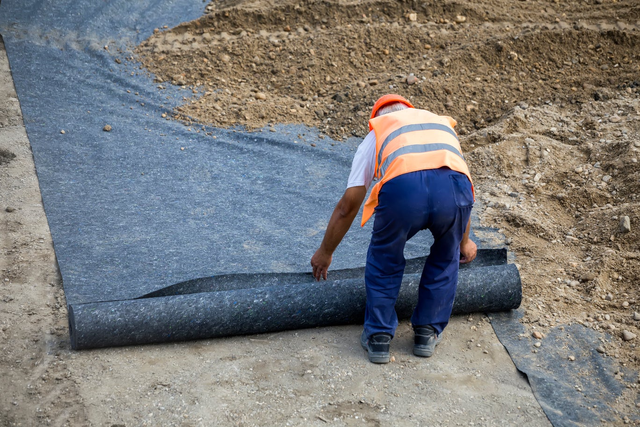 Pracownik budowlany w kamizelce odblaskowej rozwijający rolkę czarnej geowłókniny na placu budowy podczas przygotowania podłoża.