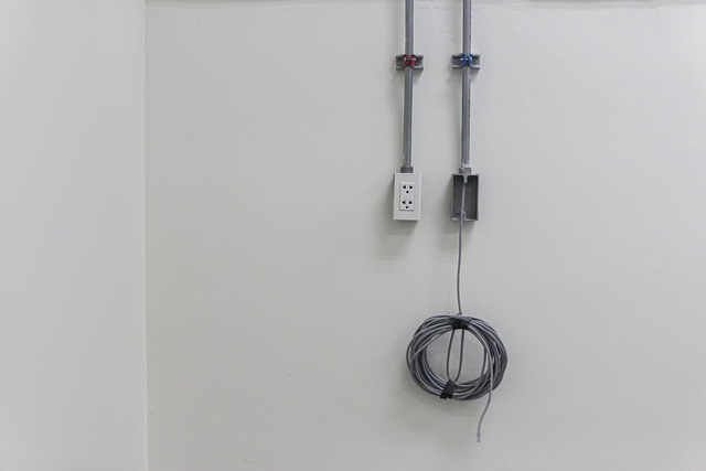 Montaż instalacji elektrycznej na białej ścianie z rurkami ochronnymi, gniazdkiem elektrycznym i zwojem kabla sieciowego.