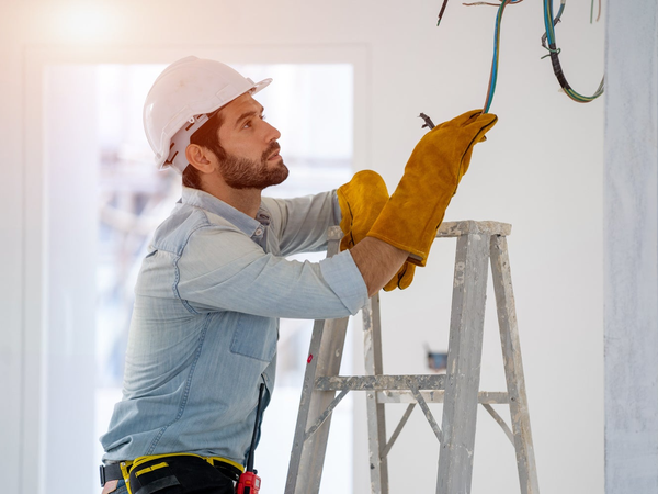 Elektryk w kasku ochronnym i rękawicach roboczych wykonujący podłączenie przewodów elektrycznych na drabinie.