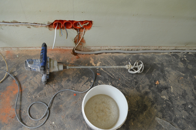 Mieszadło elektryczne z wiadrem zaprawy na podłodze podczas remontu instalacji elektrycznej z widocznymi puszkami podtynkowymi w ścianie.