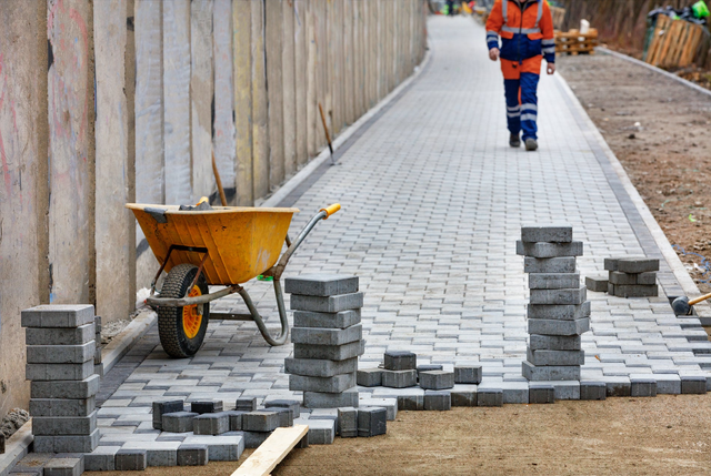 Układanie szarej kostki brukowej na chodniku przez pracownika w pomarańczowej kamizelce odblaskowej z żółtą taczką.