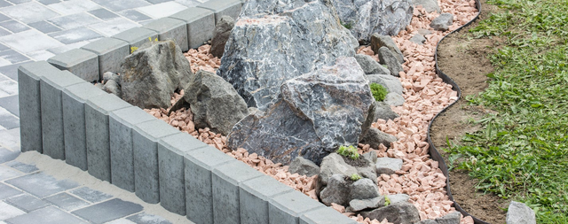 Ogrodowa kompozycja skalna z głazami granitowymi i różowym żwirem otoczona betonową palisadą przy szarej kostce brukowej.