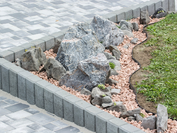 Ogrodowa kompozycja skalna z głazami granitowymi i różowym żwirem otoczona betonową palisadą przy szarej kostce brukowej.