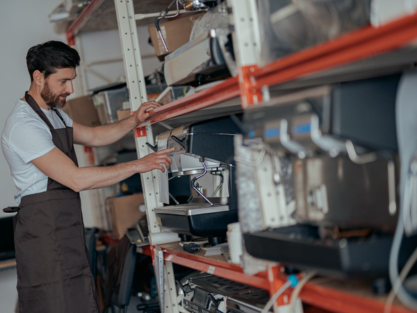 Technik serwisowy w brązowym fartuchu pobierający sprzęt elektroniczny z pomarańczowego regału magazynowego w warsztacie.