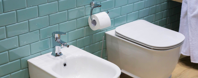 Podwieszana miska WC i bidet z chromowaną baterią na tle turkusowych płytek cegiełek z drewnianą podłogą w nowoczesnej łazience.