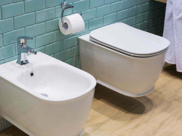 Podwieszana miska WC i bidet z chromowaną baterią na tle turkusowych płytek cegiełek z drewnianą podłogą w nowoczesnej łazience.