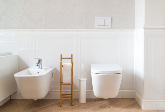 Białe podwieszane WC i bidet z bambusowym stojaczkiem na papier toaletowy i szczotką WC w jasnej łazience z parkietem w jodełkę.