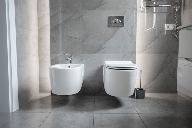 Podwieszana miska WC i bidet przy ścianie wyłożonej szarymi płytkami imitującymi marmur z chromowanym przyciskiem spłuczki podtynkowej.