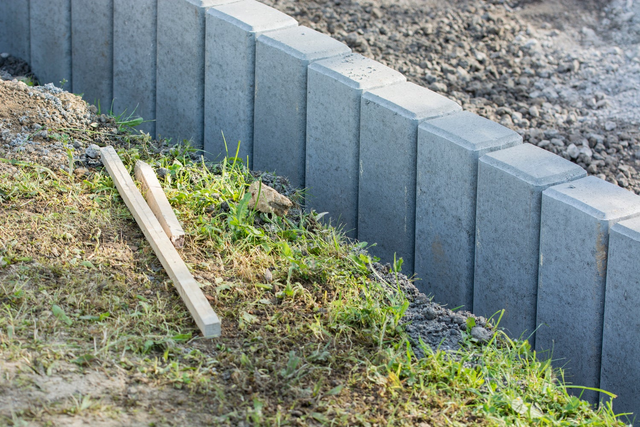 Montaż szarych palisad betonowych jako obrzeża rabaty w trakcie prac ogrodowych.