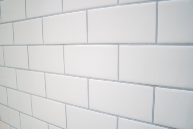 Białe płytki ceramiczne metro ułożone w cegiełkę z szarą fugą na ścianie łazienkowej.