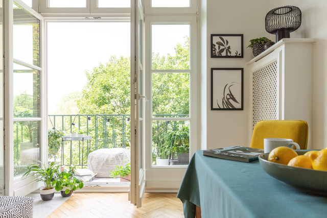 Otwarte białe drzwi balkonowe z widokiem na zielone drzewa i urządzony balkon w miejskim mieszkaniu.