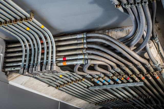 Instalacja elektryczna z szarymi rurami karbowanymi i oznaczonymi kolorowymi taśmami przewodami poprowadzonymi pod stropem budynku.