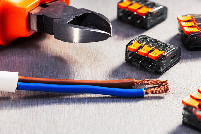 Obcęgi elektryczne, oголone przewody miedziane w niebieskiej i brązowej izolacji oraz zestaw szaro-pomarańczowych złączek sprężynowych na szarej powierzchni.