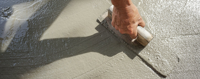 Dłoń murarza wyrównuje mokry beton pacą na posadzce podczas prac wykończeniowych.