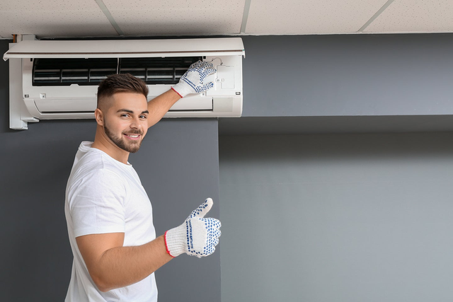 Uśmiechnięty instalator pokazuje kciuk w górę podczas montażu białego klimatyzatora ściennego na szarej ścianie.